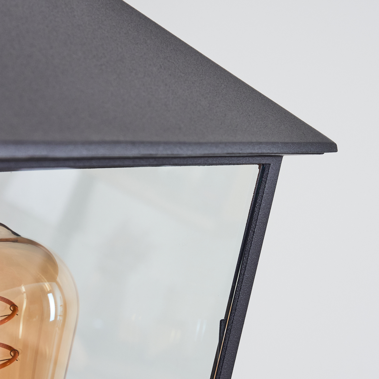 Nahaufnahme einer schwarzen Metallleuchte. Eine Glasscheibe zeigt eine Glühbirne. Der Hintergrund ist eine schlichte weiße Wand.