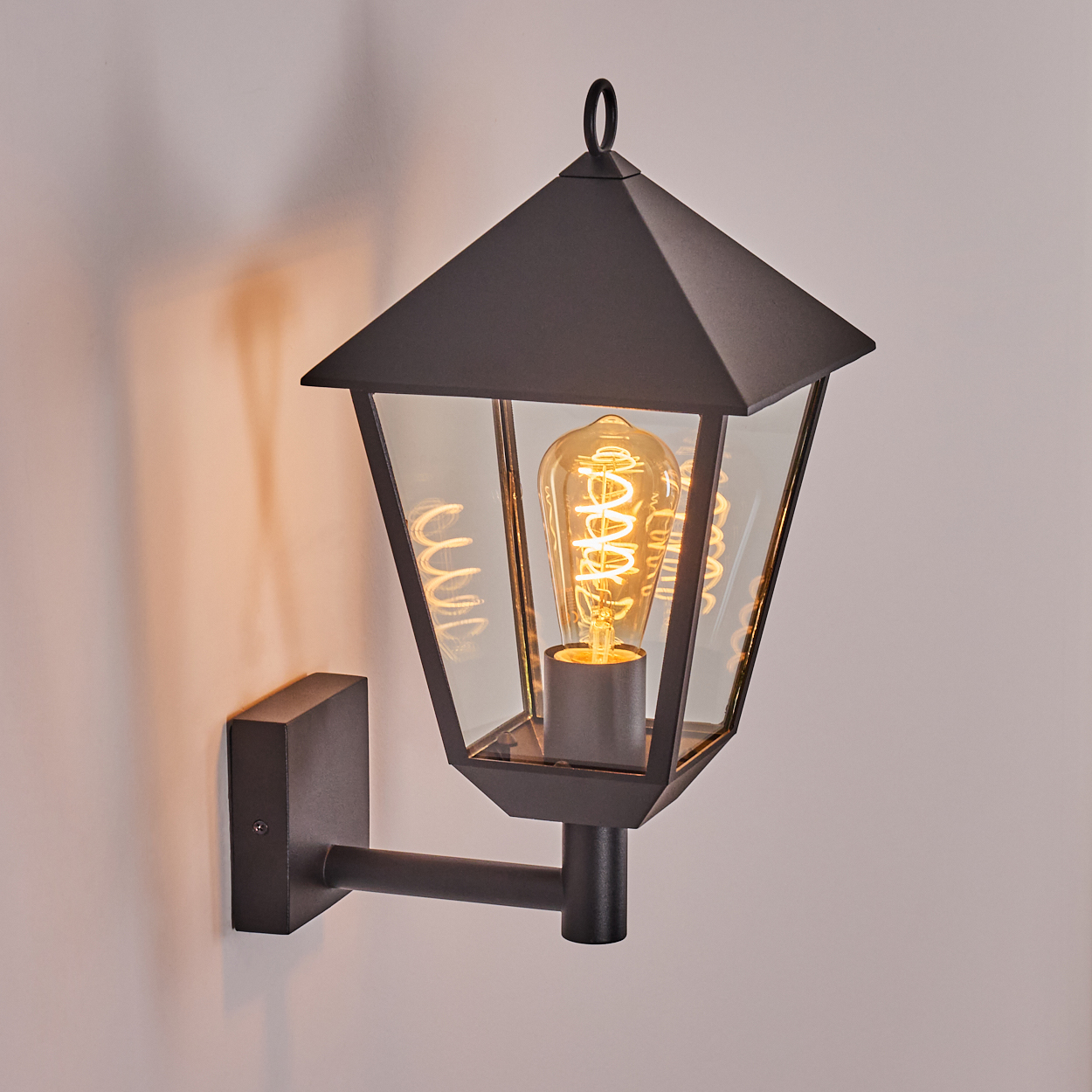 Eine schwarze, an der Wand befestigte Laterne mit Glasgehäuse und einer leuchtenden Glühbirne. Das Licht wirft einen warmen Schein an die Wand.