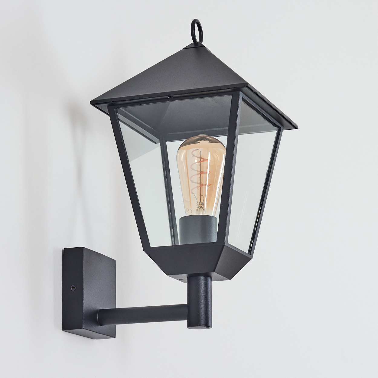 Schwarze Außenlampe mit Glasscheiben und einer Glühbirne, an einer weißen Wand montiert. Die Lampe hat ein klassisches Design.