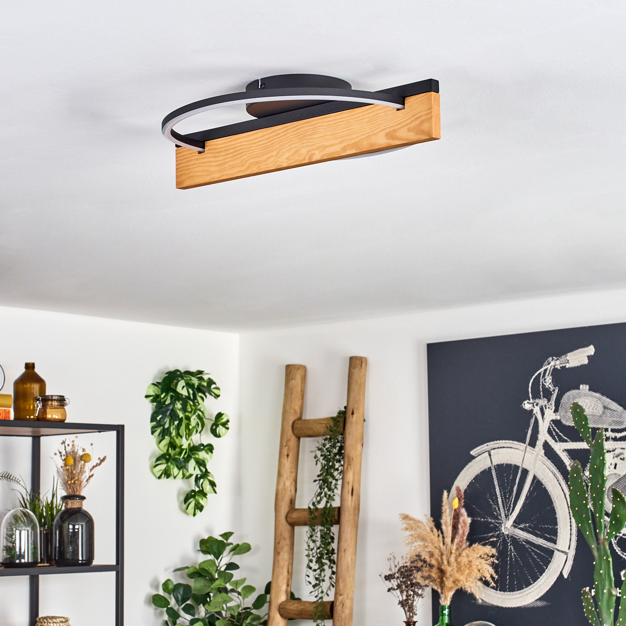 Eine moderne Deckenleuchte mit Holz- und schwarzen Akzenten. Im Hintergrund sind eine Leiter, Pflanzen und ein Fahrrad-Kunstwerk zu sehen.