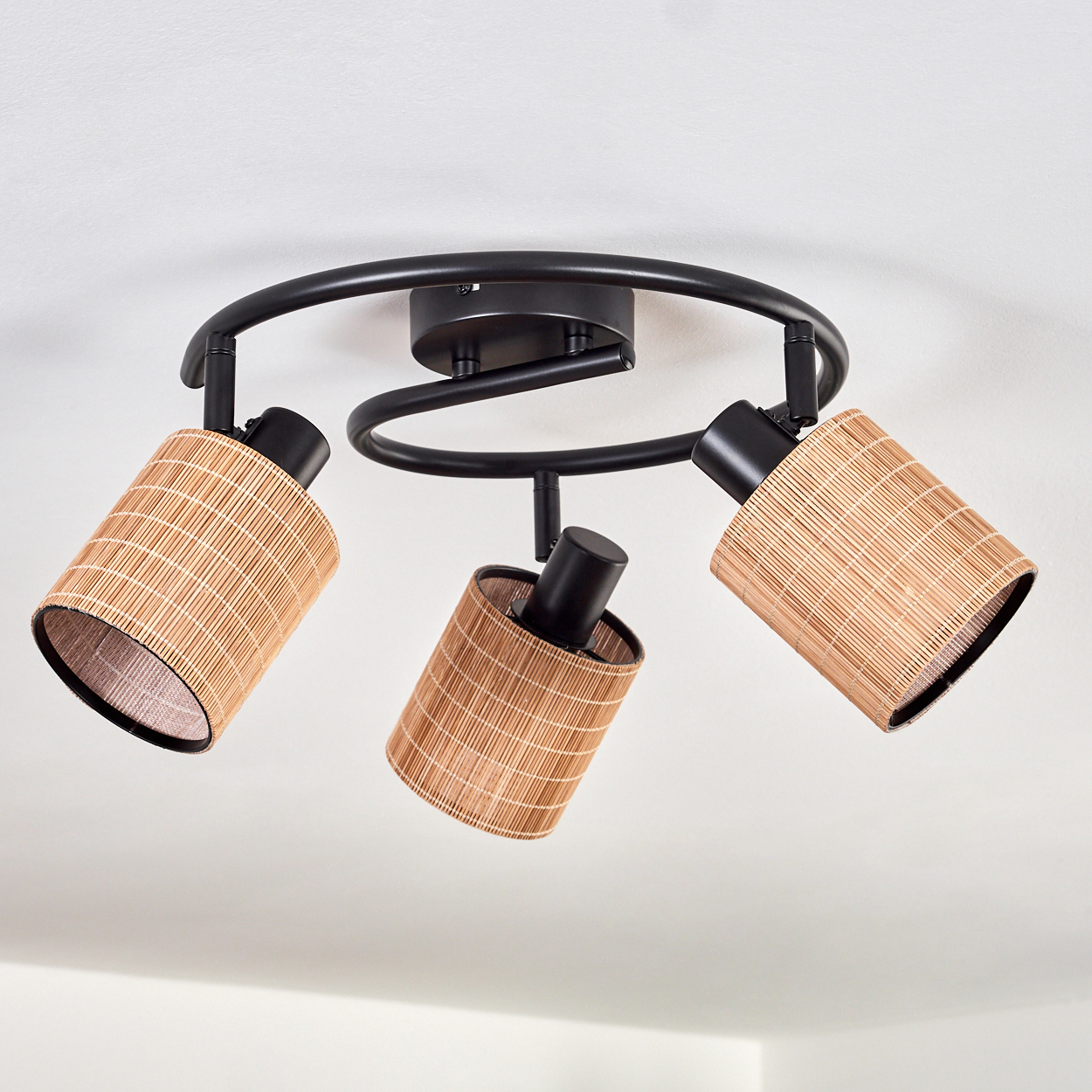 Eine Deckenleuchte mit drei Lampen. Lampen sind zylindrisch mit gewebten Schirmen. Die Leuchte ist schwarz, an einer weißen Decke befestigt.