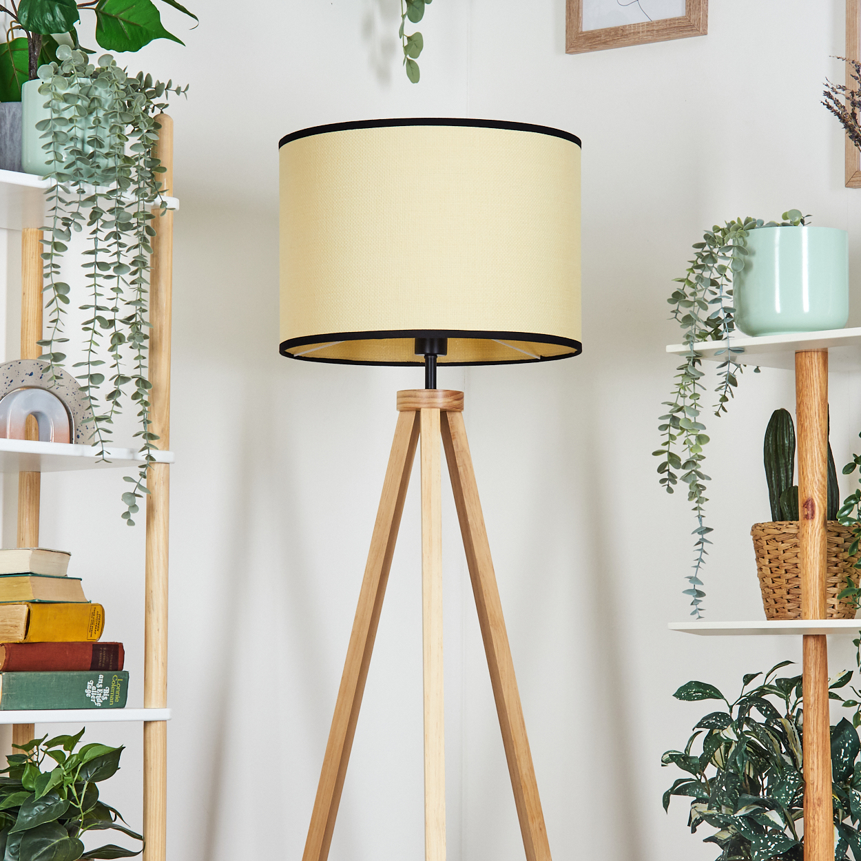 Eine Stehlampe mit beigem Lampenschirm und Holzstativfuß steht in einem Raum mit Regalen, Pflanzen und Büchern.