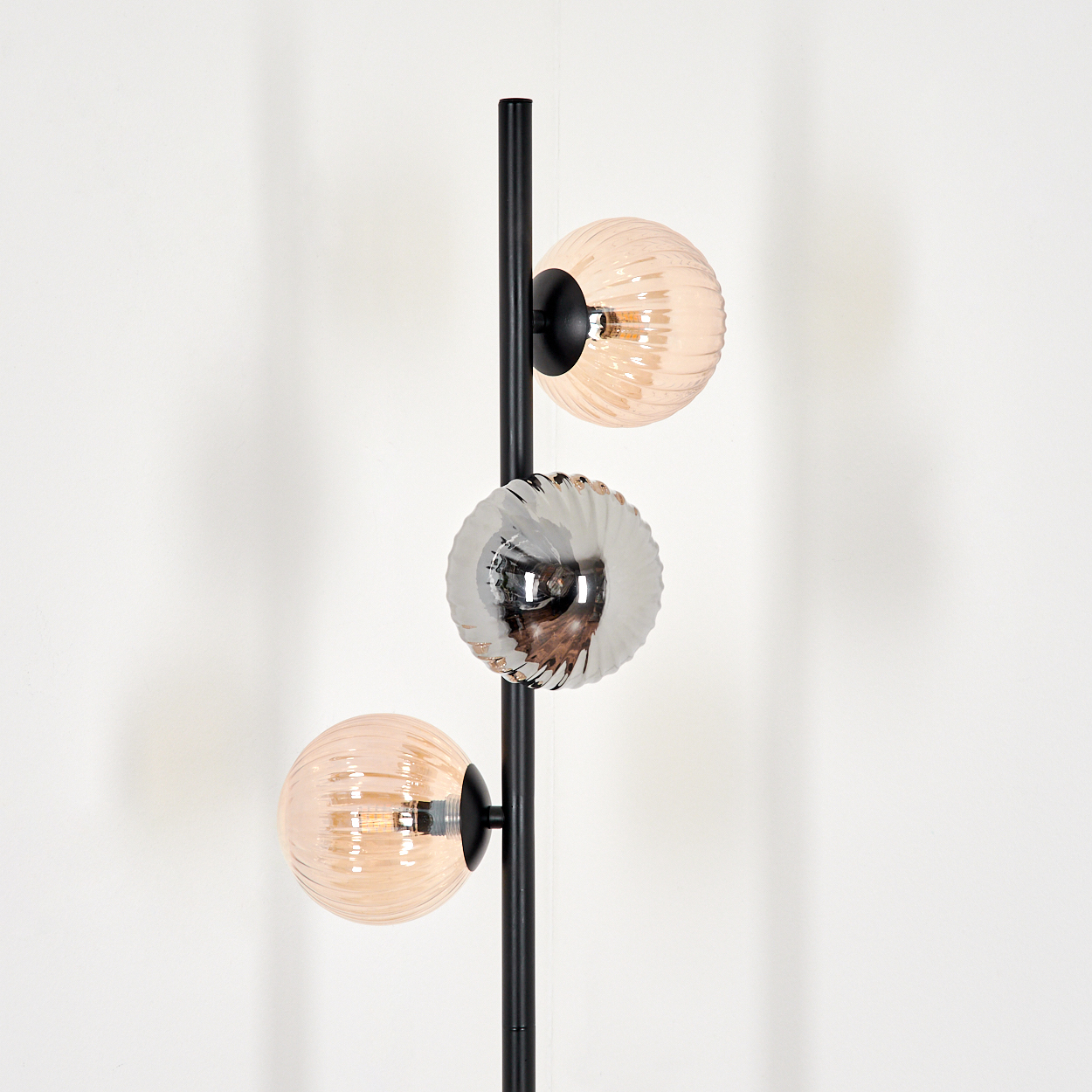 Eine schwarze Stehlampe mit drei Glaskugeln. Zwei sind hellrosa, eine ist silber. Der Hintergrund ist eine weiße Wand.