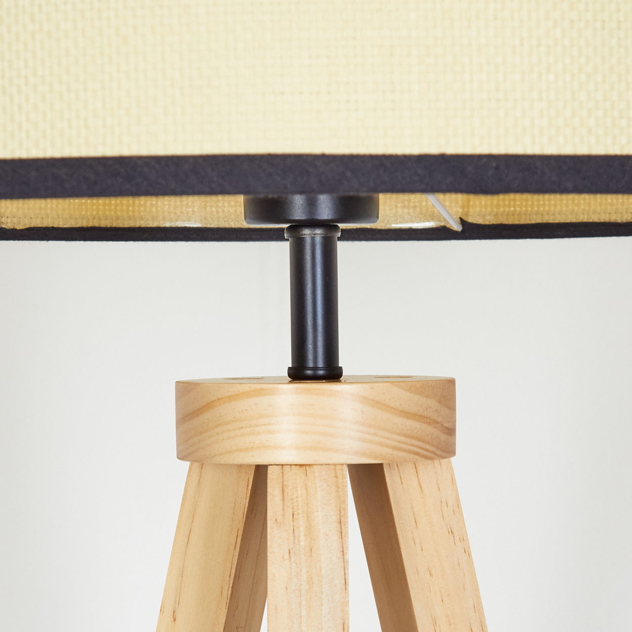 Nahaufnahme einer Holzlampe. Sie hat einen hellbeigen Lampenschirm, einen schwarzen Metallschaft und einen hölzernen Dreibein-Sockel vor weißem Hintergrund.