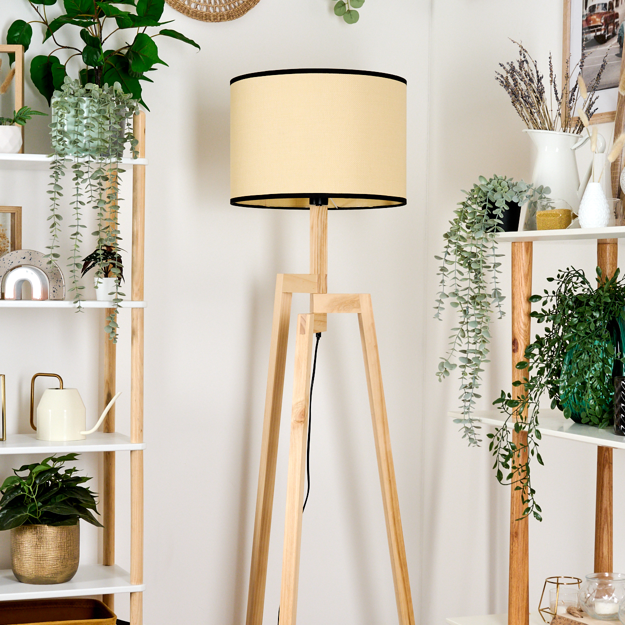 Eine Stehlampe aus Holz mit beigem Lampenschirm ist von Regalen mit Pflanzen, einer Gießkanne und Dekorationsartikeln umgeben.
