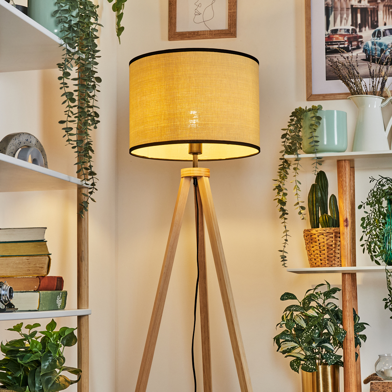 Eine hölzerne Stehlampe mit beigem Lampenschirm steht in einem Raum mit Pflanzen, Büchern und Regalen. Wandkunst sichtbar.