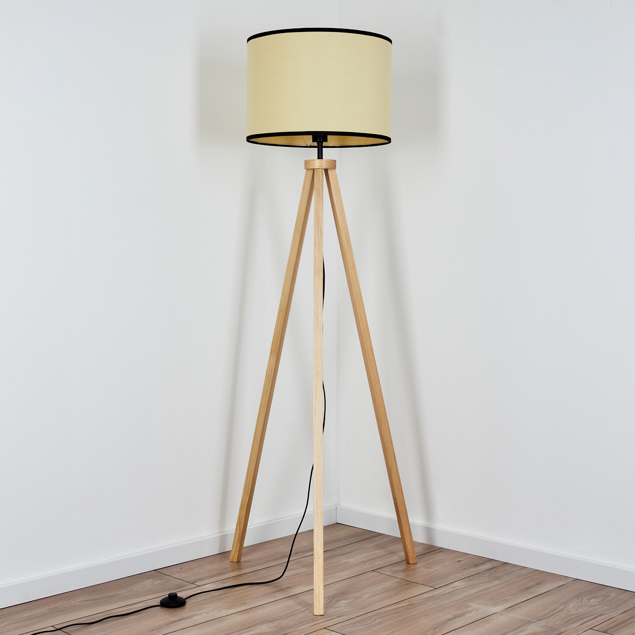 Eine hölzerne Stativ-Stehlampe mit beigem Lampenschirm und schwarzem Rand, die in einer Ecke steht. Der Boden ist aus Holz und die Wände sind weiß.