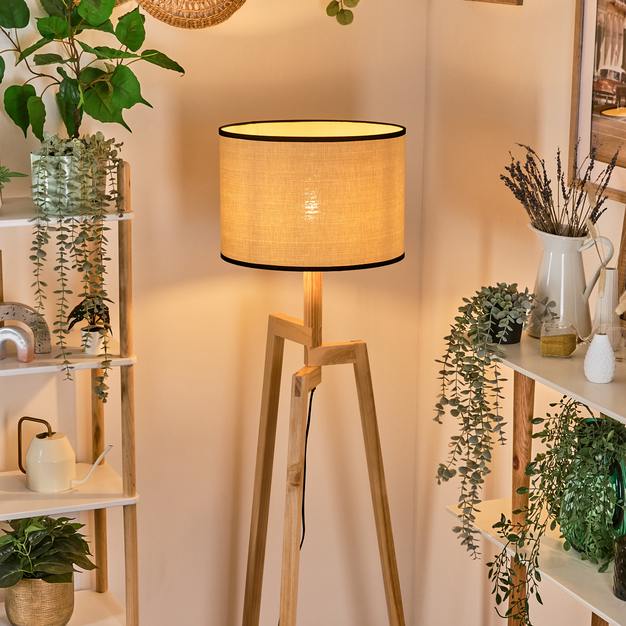 Eine Stehlampe aus Holz mit beigem Schirm steht in einer Ecke. Regale mit Pflanzen befinden sich auf beiden Seiten. Eine weiße Gießkanne ist sichtbar.