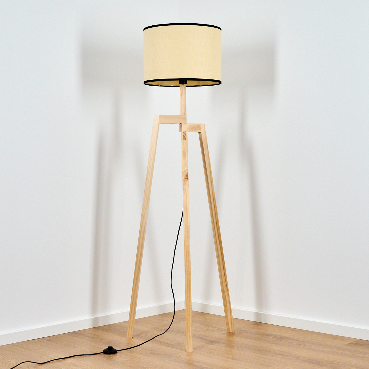 Eine hölzerne Stehlampe mit beigem Lampenschirm und schwarzem Rand steht in einer Ecke auf einem Holzboden.
