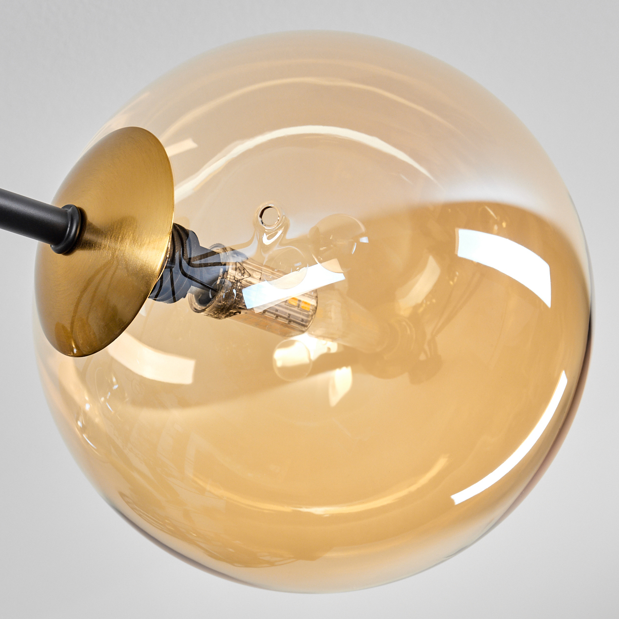 Nahaufnahme einer modernen Lampe mit rundem, bernsteinfarbenem Glasschirm. Eine goldene Metallscheibe und ein schwarzer Arm tragen den Schirm. Der Hintergrund ist weiß.
