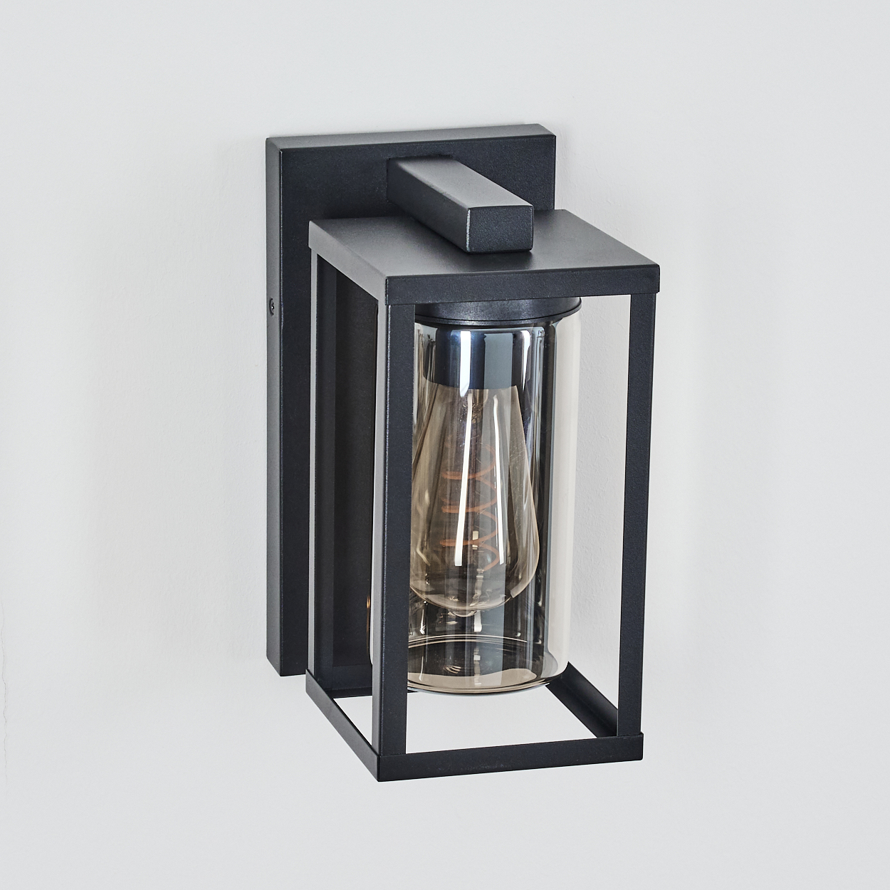 Eine schwarze Außenwandleuchte. Sie hat einen rechteckigen Rahmen mit einem Glaszylinder, der eine Glühbirne enthält. An einer weißen Wand montiert.