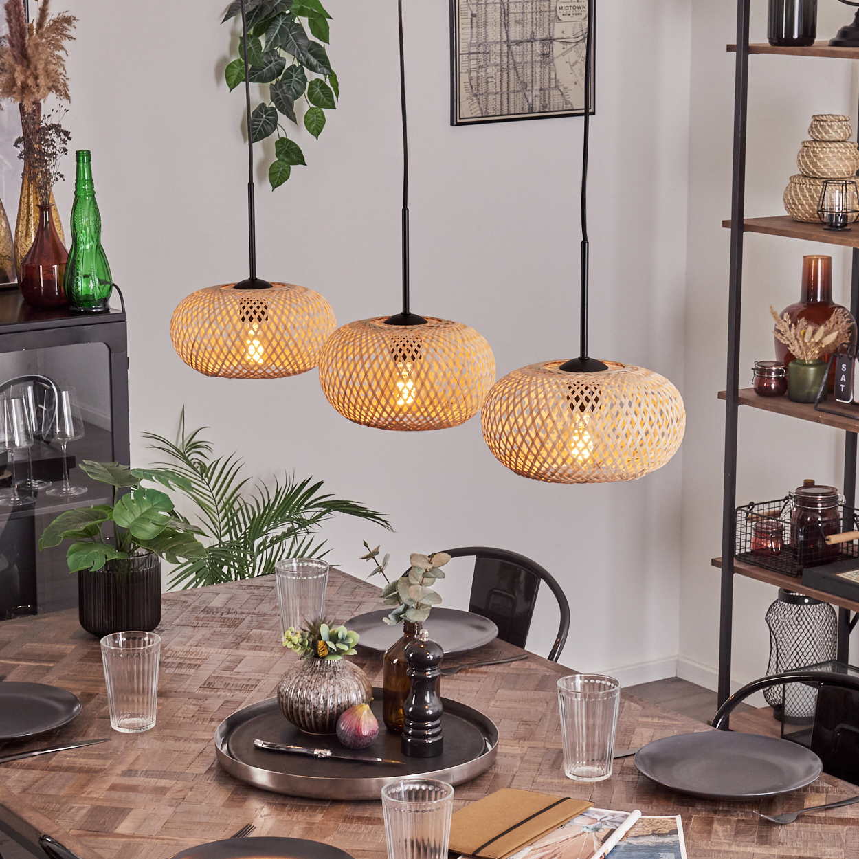 Ein gedeckter Esstisch mit drei gewebten Lampen darüber. Teller, Gläser und Dekoration stehen auf dem Tisch. Im Hintergrund ein Regal.