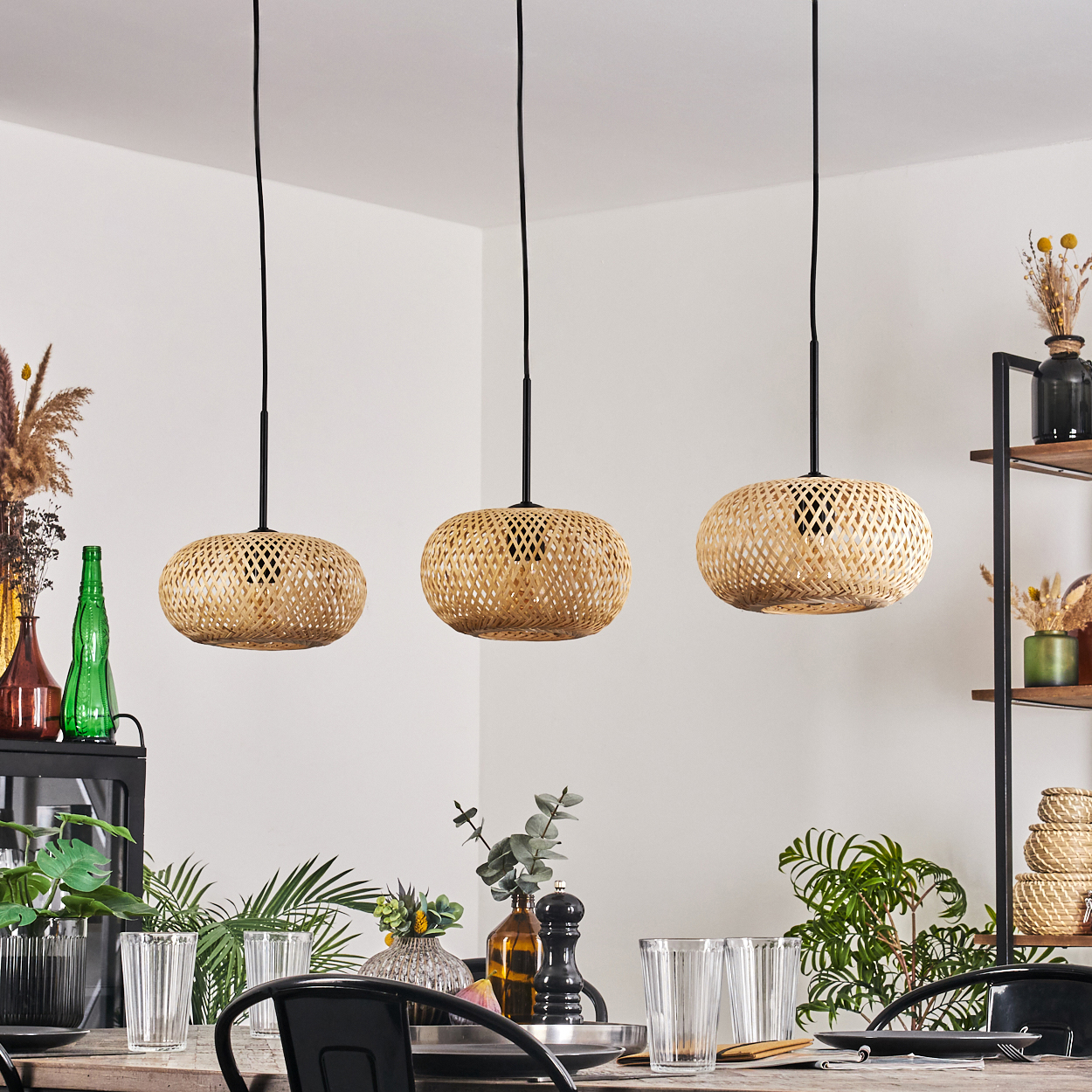Drei gewebte Lampen hängen über einem Tisch, der für eine Mahlzeit gedeckt ist. Im Hintergrund grüne Pflanzen und Vasen.