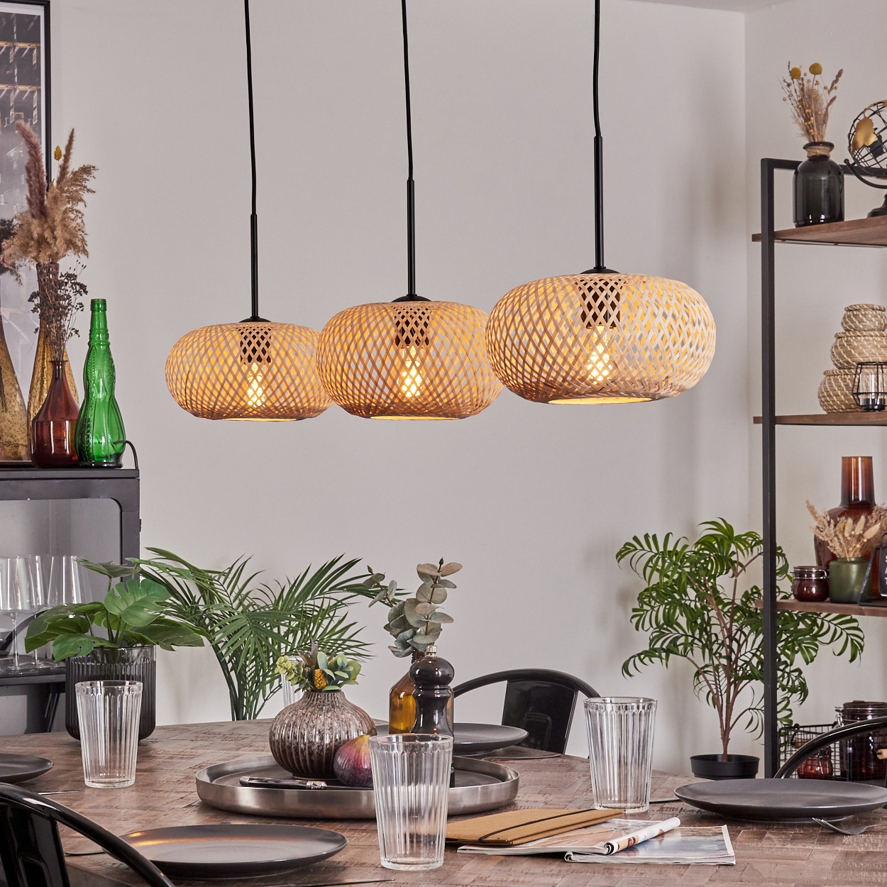Drei Bambuslampen hängen über einem Holzesstisch mit Tellern, Gläsern und einem Mittelstück. Pflanzen und Regale im Hintergrund.