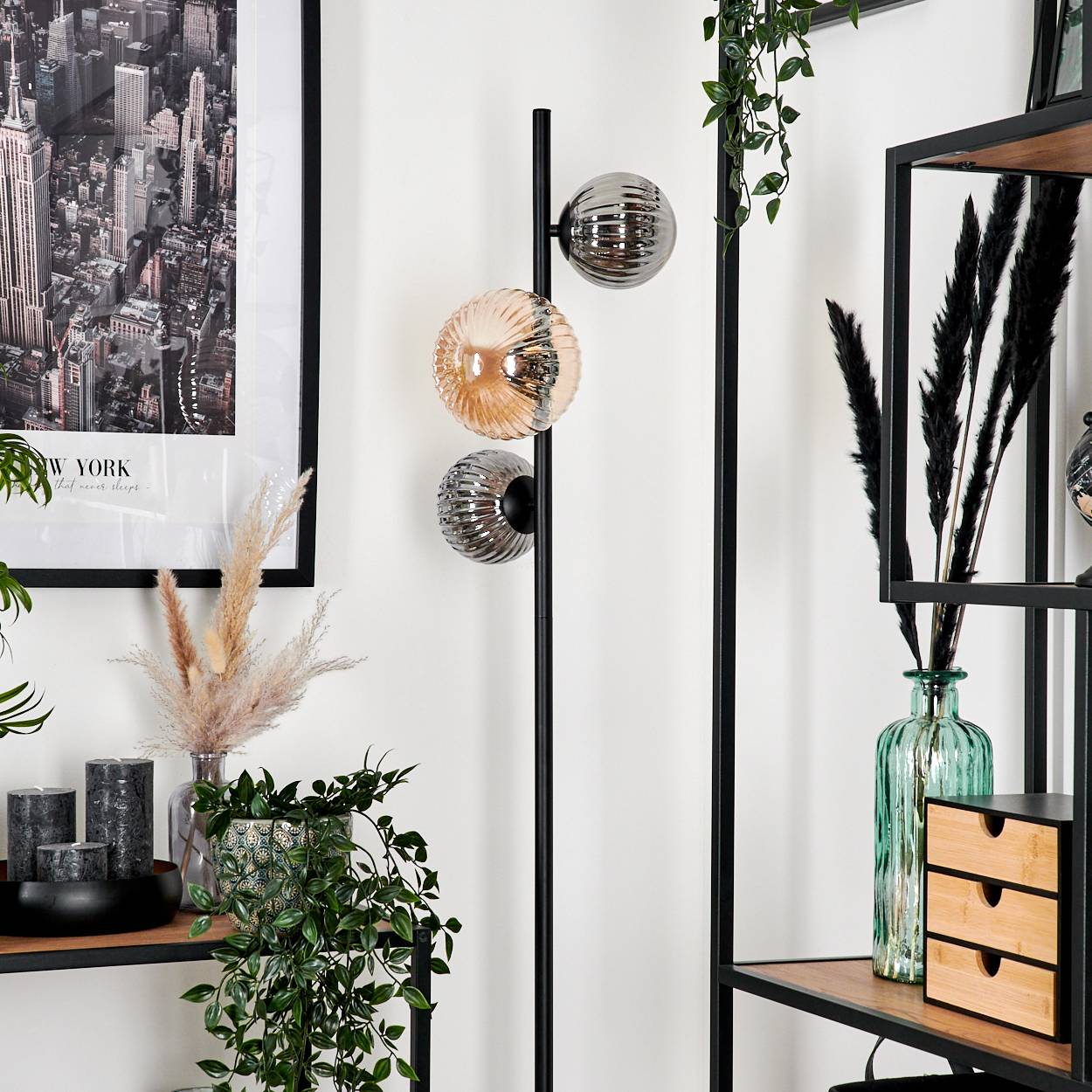 Ein Rauminnenraum zeigt eine schwarze Stehlampe mit drei Kugelleuchten. Regale mit Pflanzen, Dekoration und einem NYC-Druck sind sichtbar.