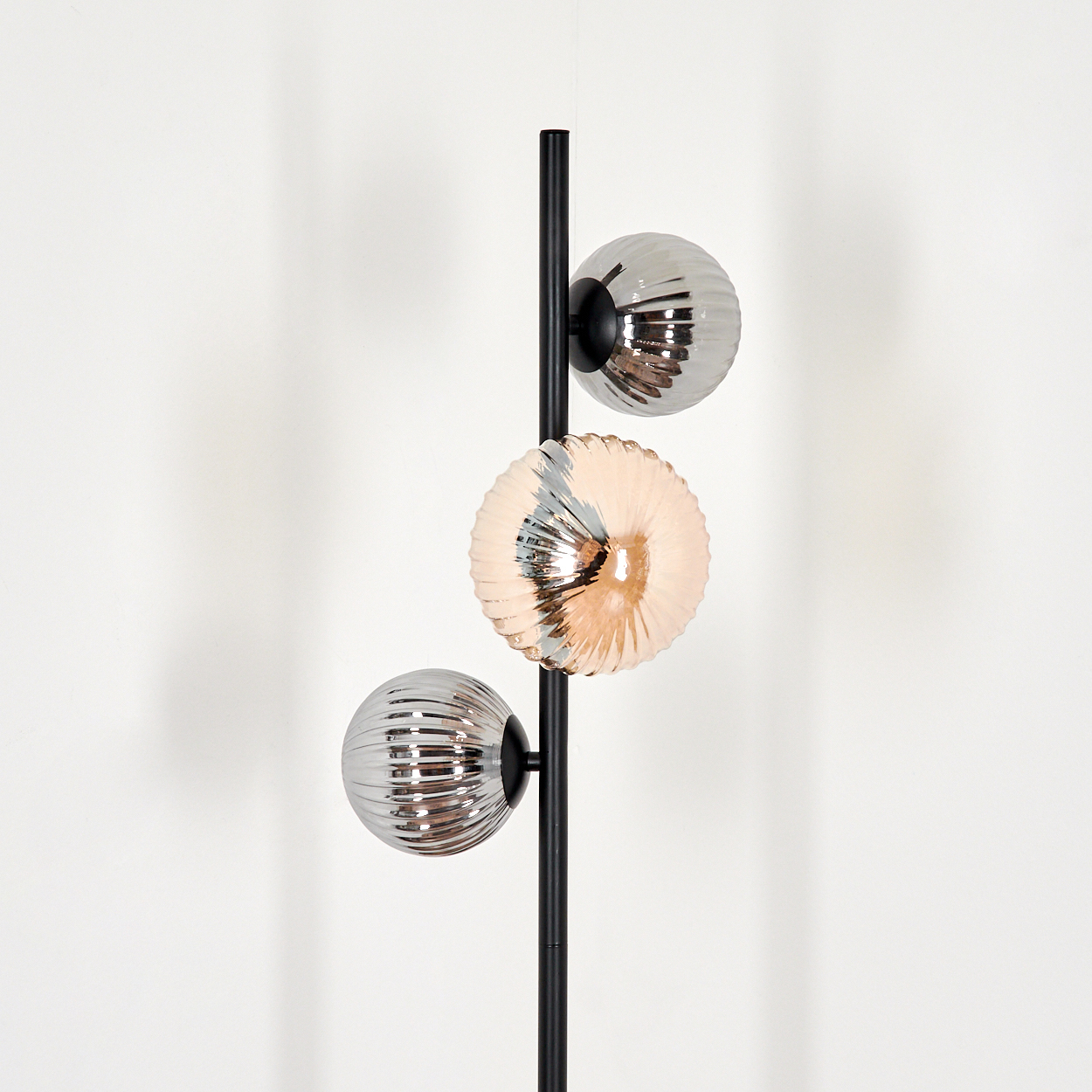 Eine schwarze Stehlampe mit drei kugelförmigen, gerippten Glasschirmen in Silber- und Pfirsichfarben. Der Hintergrund ist eine weiße Wand.