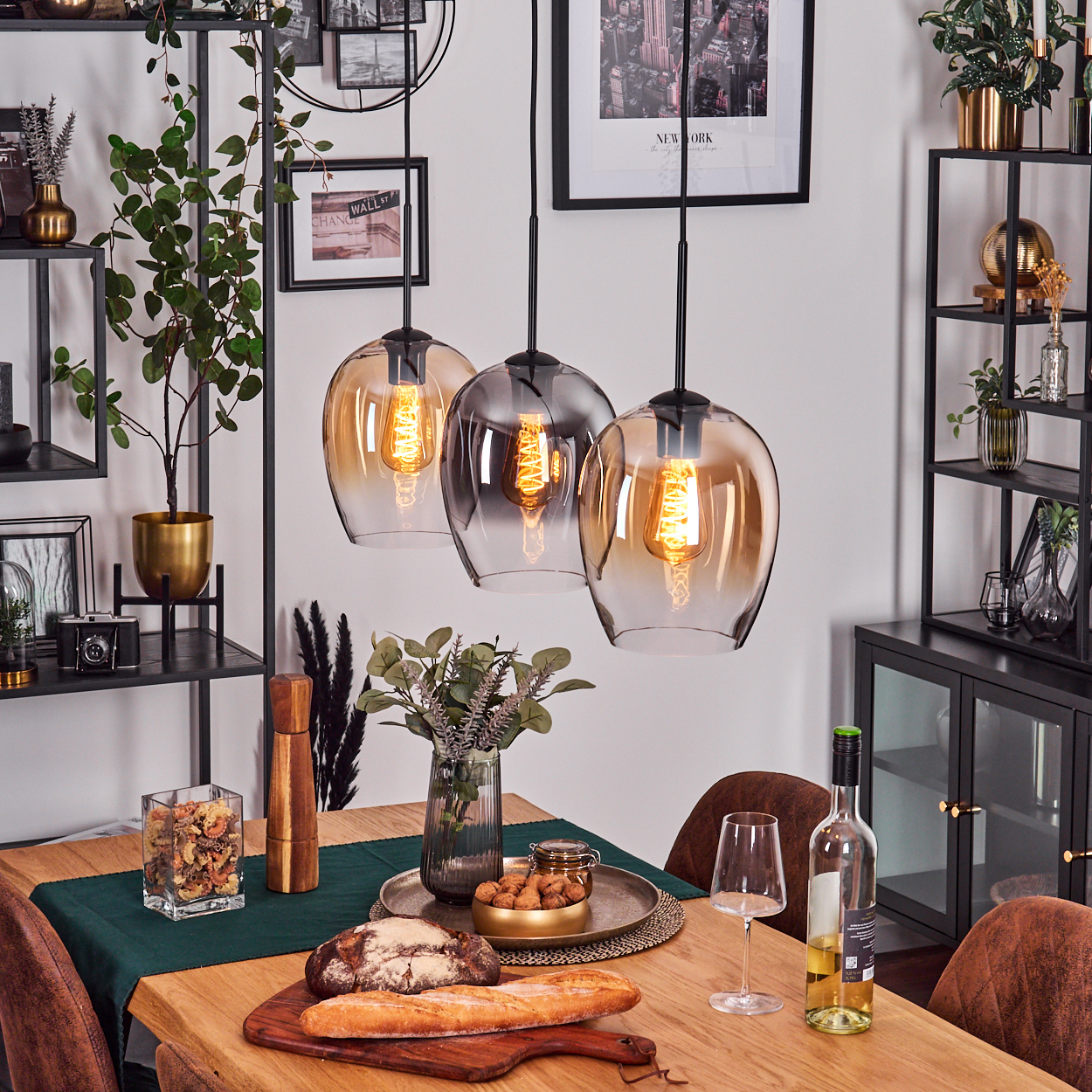 Esszimmer mit drei Pendelleuchten, Tisch mit Speisen und Wein sowie dekorativen Regalen mit Pflanzen und Dekoration.
