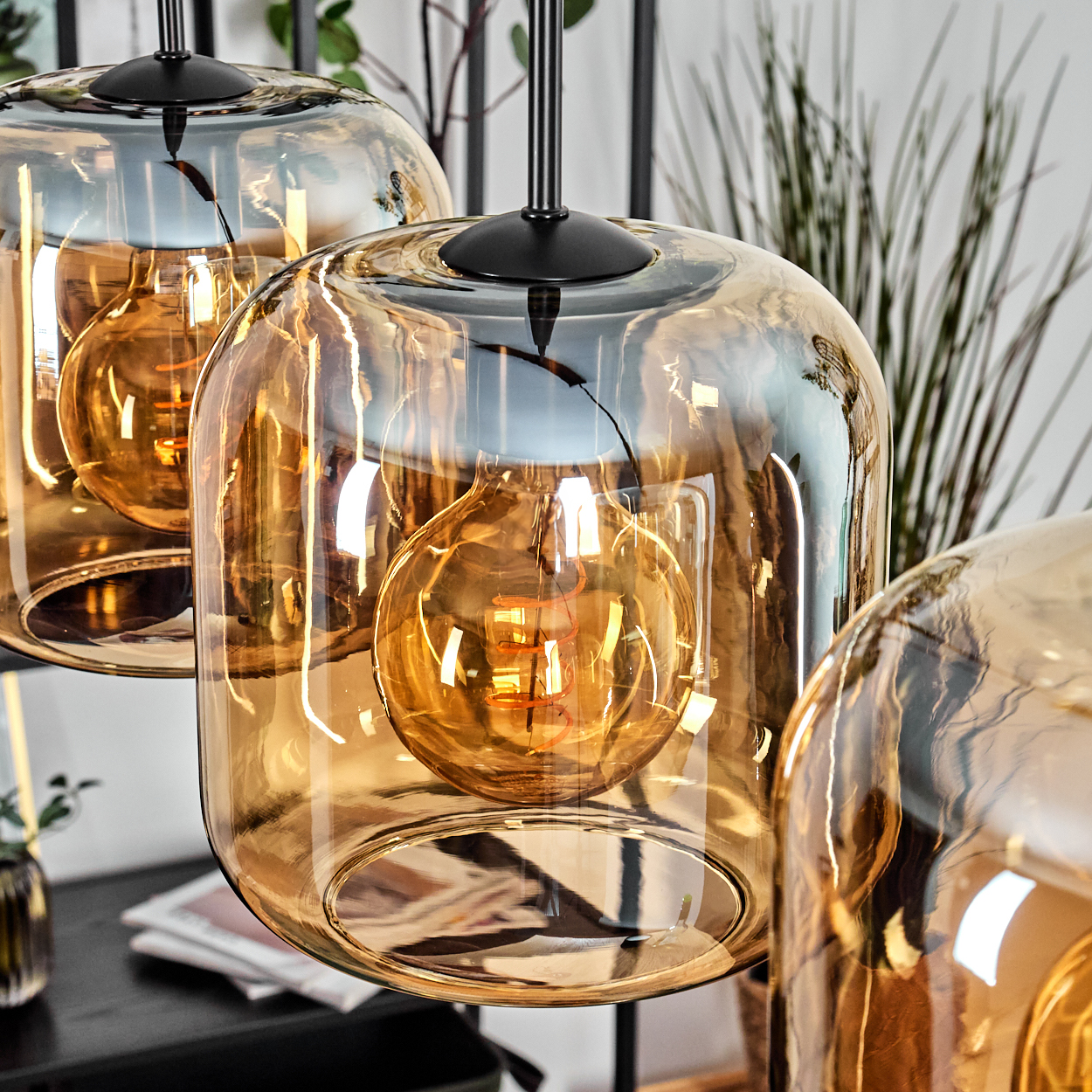 Nahaufnahme von drei Pendelleuchten mit bernsteinfarbenen Glasschirmen und Edison-Glühbirnen, schwarzen Stielen. Der Hintergrund hat Pflanzen und einen Tisch.