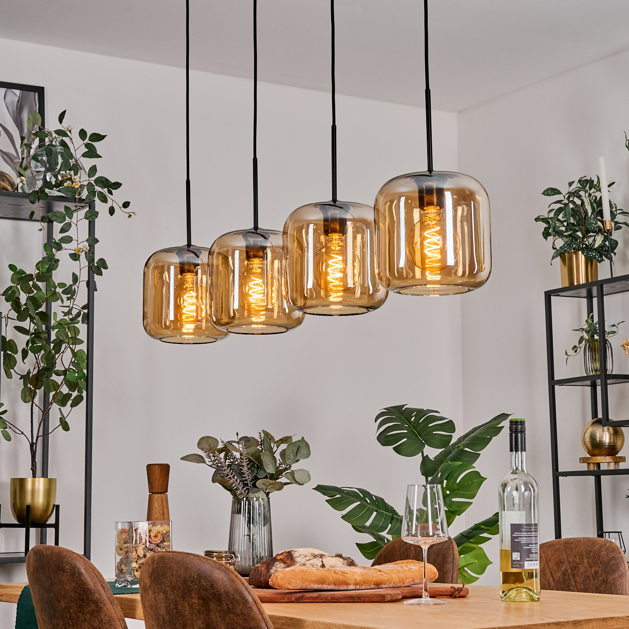 Esszimmer. Vier bernsteinfarbene Pendelleuchten hängen über einem Holztisch mit Speisen und Wein. Pflanzen und Regale im Hintergrund.