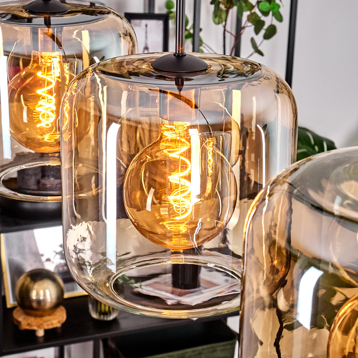 Nahaufnahme von drei Pendelleuchten mit bernsteinfarbenen Glasschirmen und leuchtenden Wendelglühbirnen. Hintergrund: Deko, Pflanzen und ein Regal.