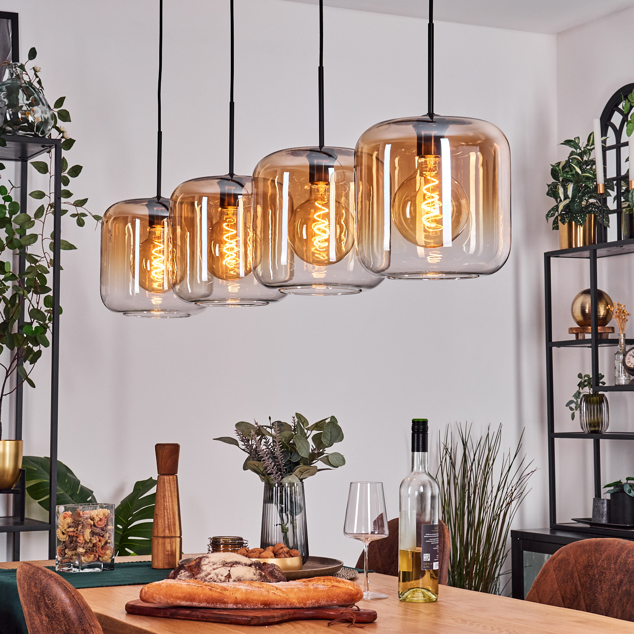 Esszimmer mit Hängelampen, einem Tisch mit Essen, Wein und Gläsern. Regale mit Pflanzen. Warme Beleuchtung.
