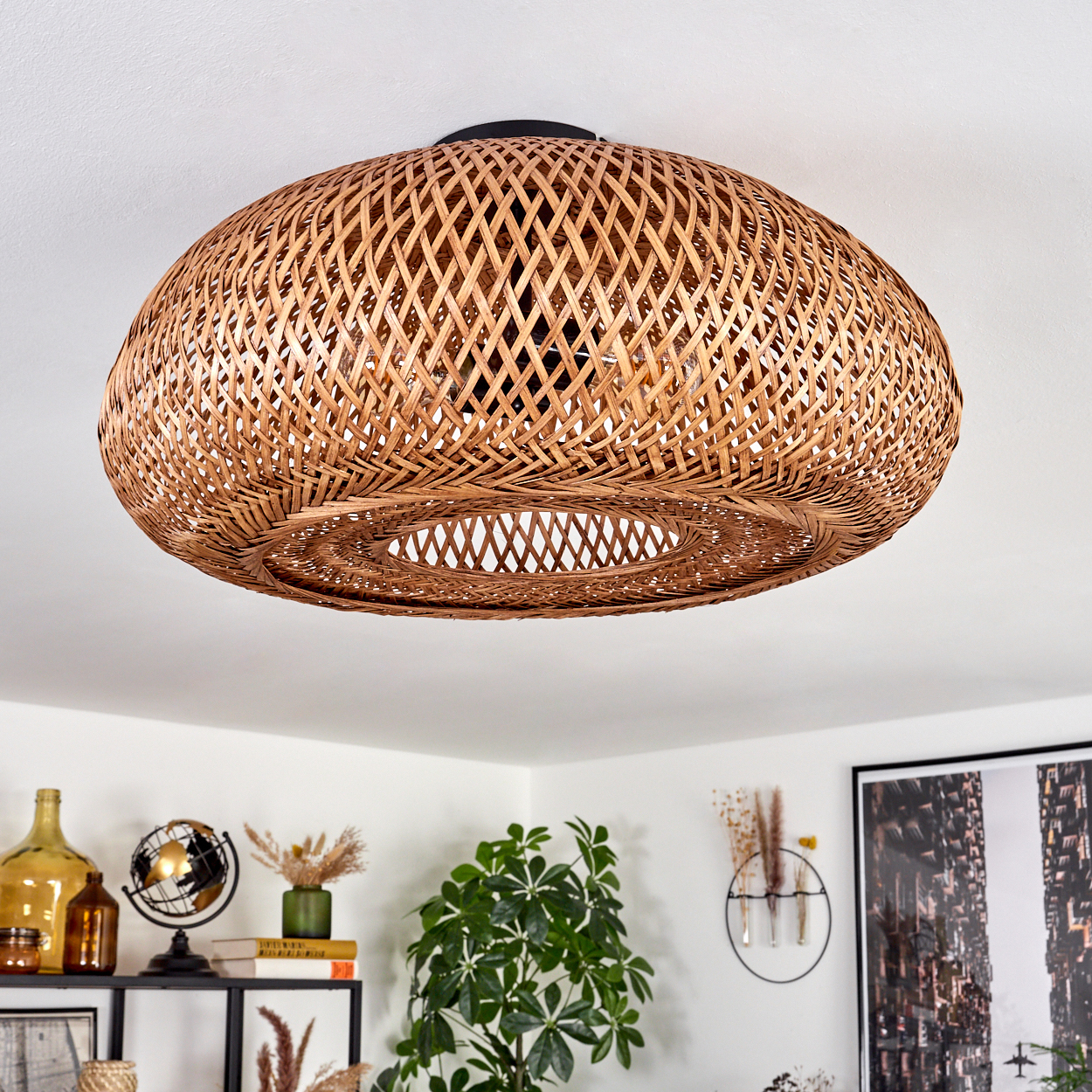Eine gewebte Deckenlampe aus Weide hängt. Darunter befindet sich ein Regal mit Dekoration. Eine Pflanze und ein gerahmtes Stadtbild sind sichtbar.
