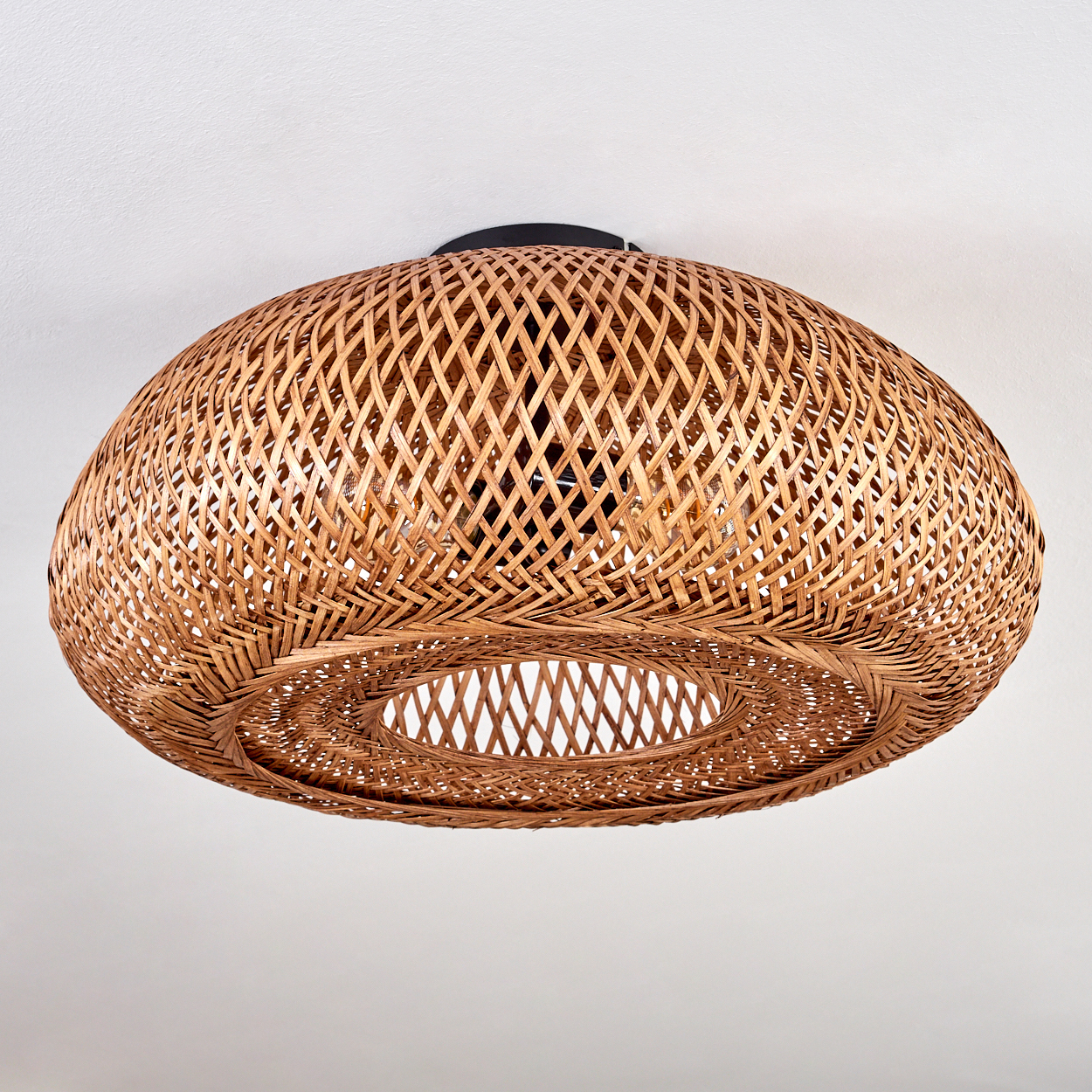 Eine geflochtene Pendelleuchte aus Rattan hängt von einer weißen Decke. Das Licht ist braun und hat eine ovale Form mit einem Loch in der Mitte.