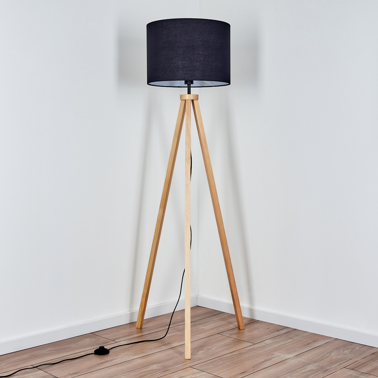 Eine Stehlampe mit schwarzem Lampenschirm und hölzernen Stativbeinen steht in einer Ecke. Der Boden ist aus Holz, die Wände sind weiß.