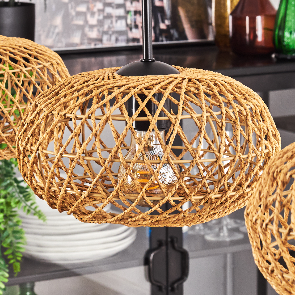 Geflochtene Pendelleuchten aus Rattan mit freiliegenden Glühbirnen. Der Hintergrund zeigt ein verschwommenes Regal mit Gläsern und Dekoration.