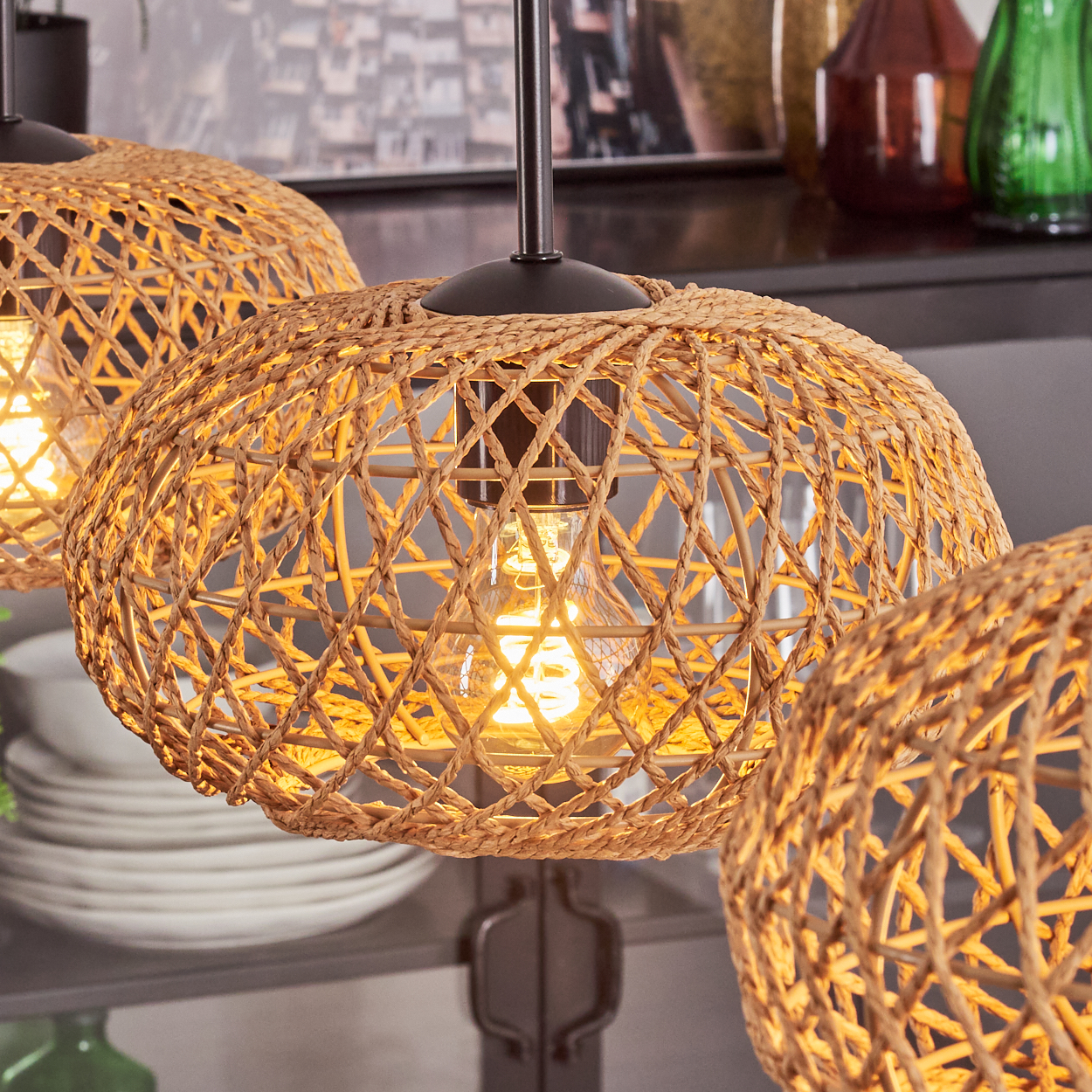Drei gewebte Lampen hängen, jede mit einer leuchtenden Glühbirne. Der Hintergrund zeigt Regale mit Flaschen und Tellern. Warme Beleuchtung.