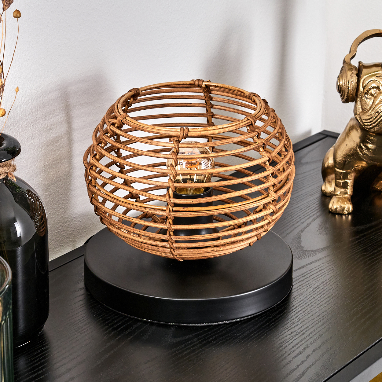 Eine gewebte Rattanlampe mit Glühbirne auf schwarzem Sockel steht auf einem schwarzen Tisch. Eine goldene Bulldoggen-Dekoration befindet sich im Hintergrund.