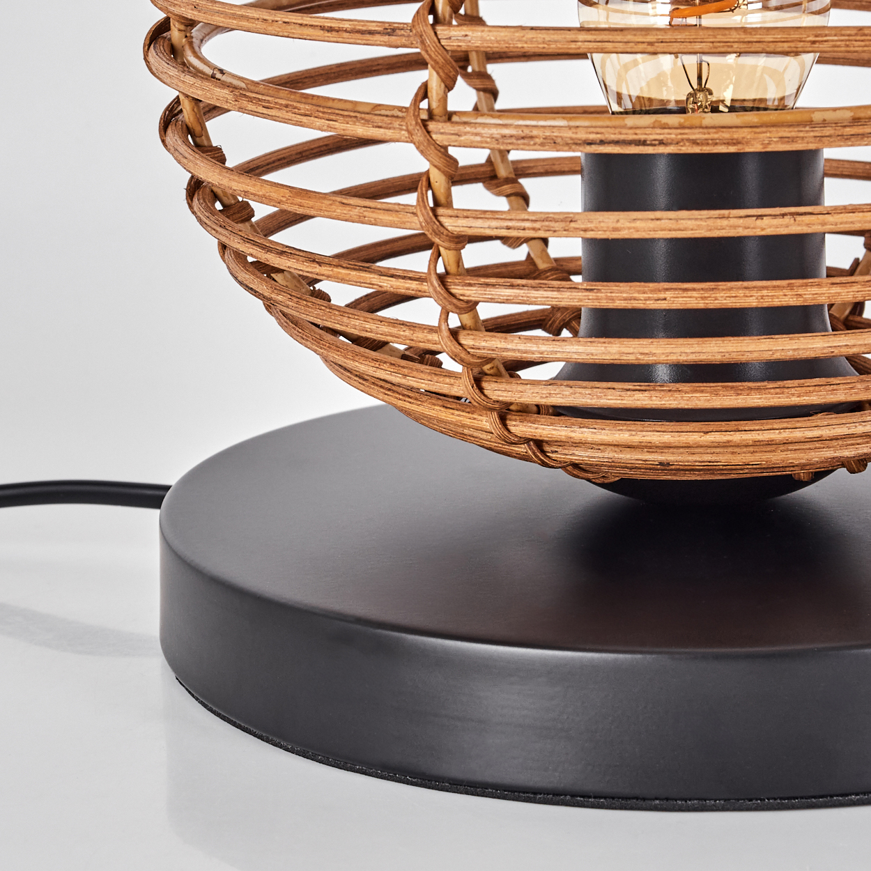 Nahaufnahme einer Lampe. Sie hat einen Schirm aus geflochtenem Rattan, einen schwarzen Sockel und eine sichtbare Glühbirne. Ein schwarzes Kabel.