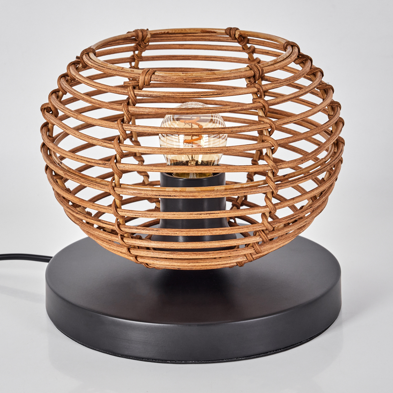 Eine kugelförmige Rattanlampe mit einer Glühbirne im Inneren steht auf einem schwarzen Sockel. Ein schwarzes Kabel geht vom Sockel aus.