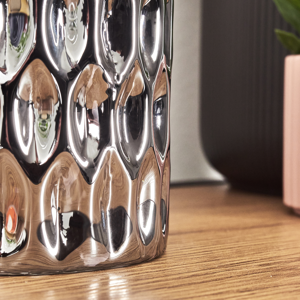 Nahaufnahme einer dekorativen silbernen Vase mit strukturierter Oberfläche. Ein Holztisch, ein dunkelgrauer Topf und ein rosa Topf sind sichtbar.
