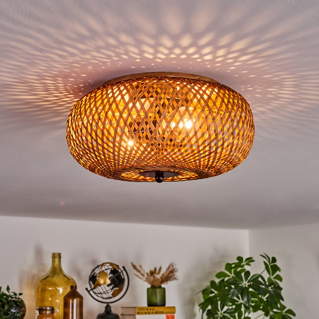 Eine Deckenlampe aus geflochtenem Bambus wirft ein gemustertes Licht an die Decke. Darunter zeigt ein Regal Dekoration, darunter Glasflaschen und Pflanzen.