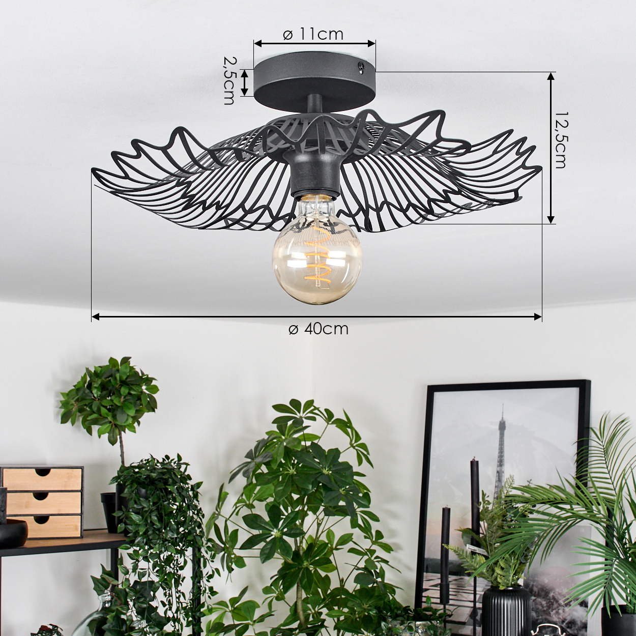 Eine schwarze Deckenlampe mit flügelartigem Design und einer Glühbirne hängt über grünen Pflanzen und einem gerahmten Bild. Maße werden angezeigt.