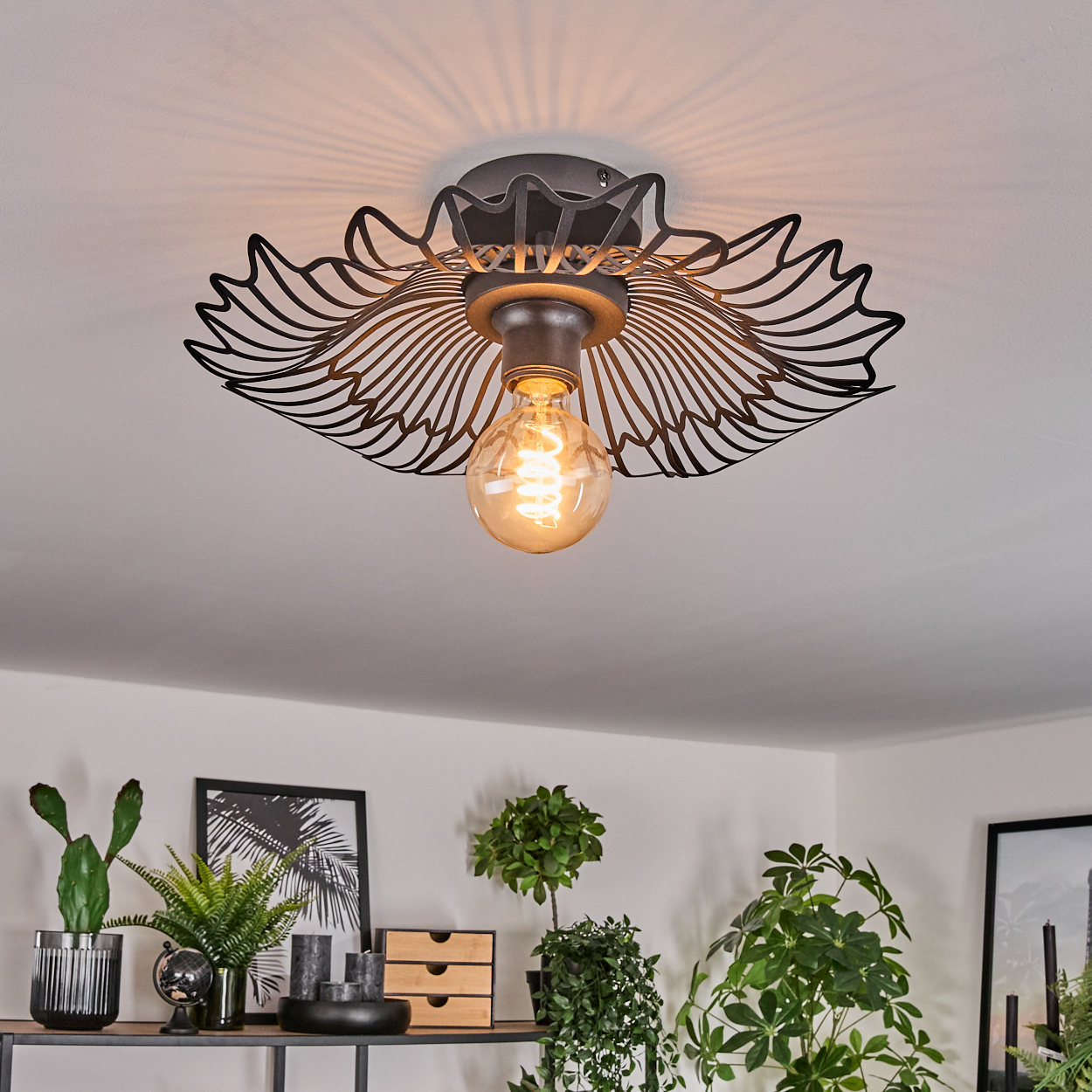 Eine Deckenleuchte mit dekorativem schwarzem Metallschirm und warm getönter Glühbirne. Pflanzen und gerahmte Kunstwerke sind unten sichtbar.