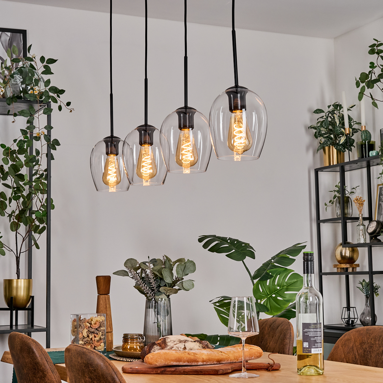 Esszimmer-Szene mit Hängelampen, Tisch mit Essen, Wein und Pflanzen auf Regalen. Braune Stühle und weiße Wände sind vorhanden.