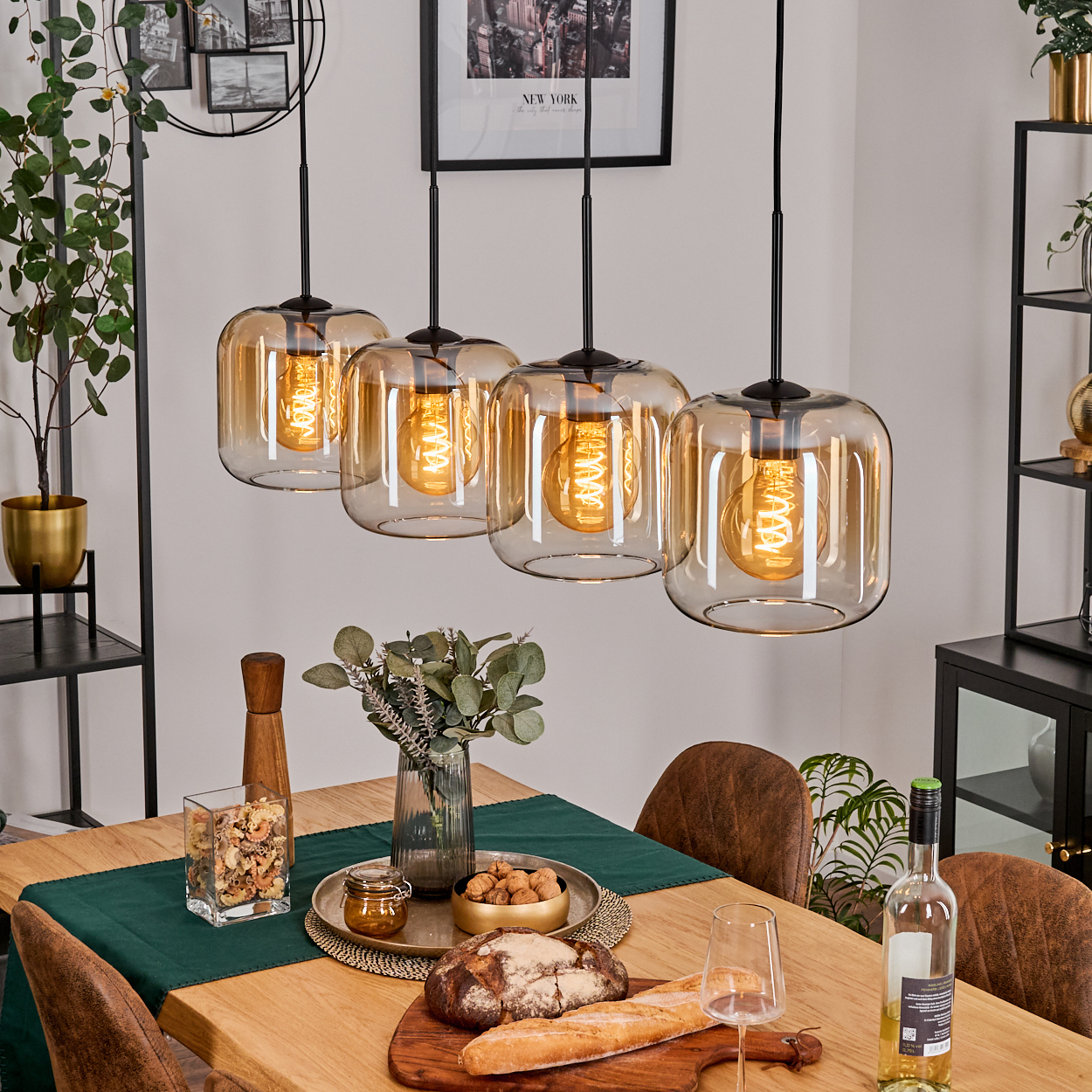 Esszimmer mit vier Hängelampen, einem Holztisch mit Speisen und Getränken. Hintergrund mit Pflanzen und Kunst.