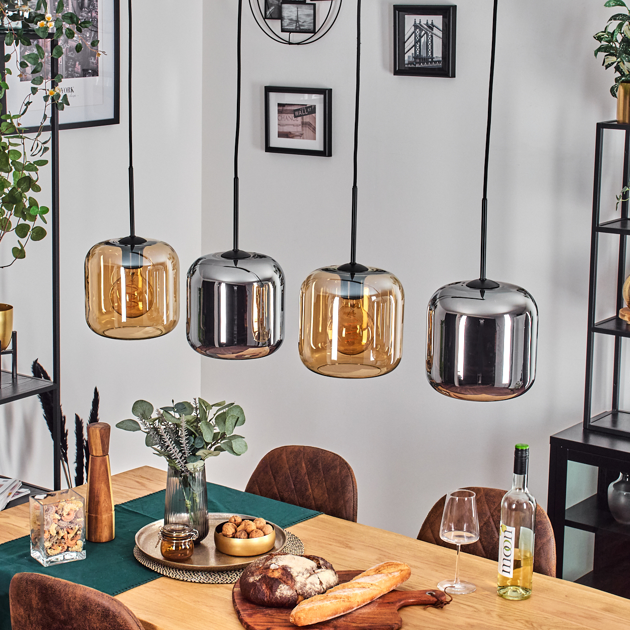 Esszimmer mit vier Hängelampen, Holztisch mit Essen und Wein, gerahmten Bildern an der weißen Wand und einem schwarzen Regal.