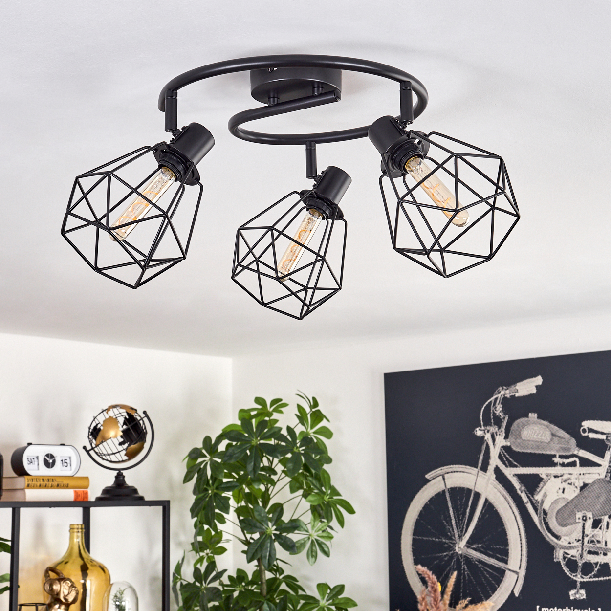 Schwarze Deckenleuchte mit drei geometrischen, käfigartigen Schirmen. Im Hintergrund ein Fahrradkunstwerk, Pflanzen und Dekorationsartikel.