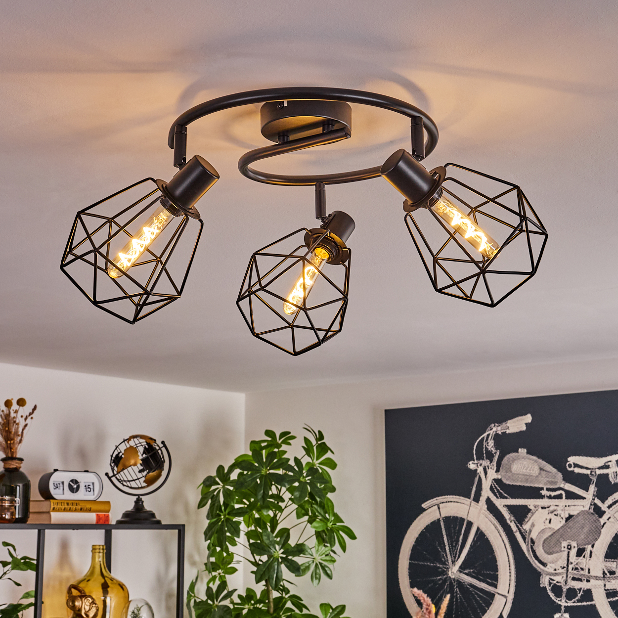 Eine schwarze Deckenlampe mit drei käfigartigen Schirmen, jeweils mit einer Glühbirne. Im Hintergrund eine Pflanze, Dekoration und ein Fahrradkunstwerk.