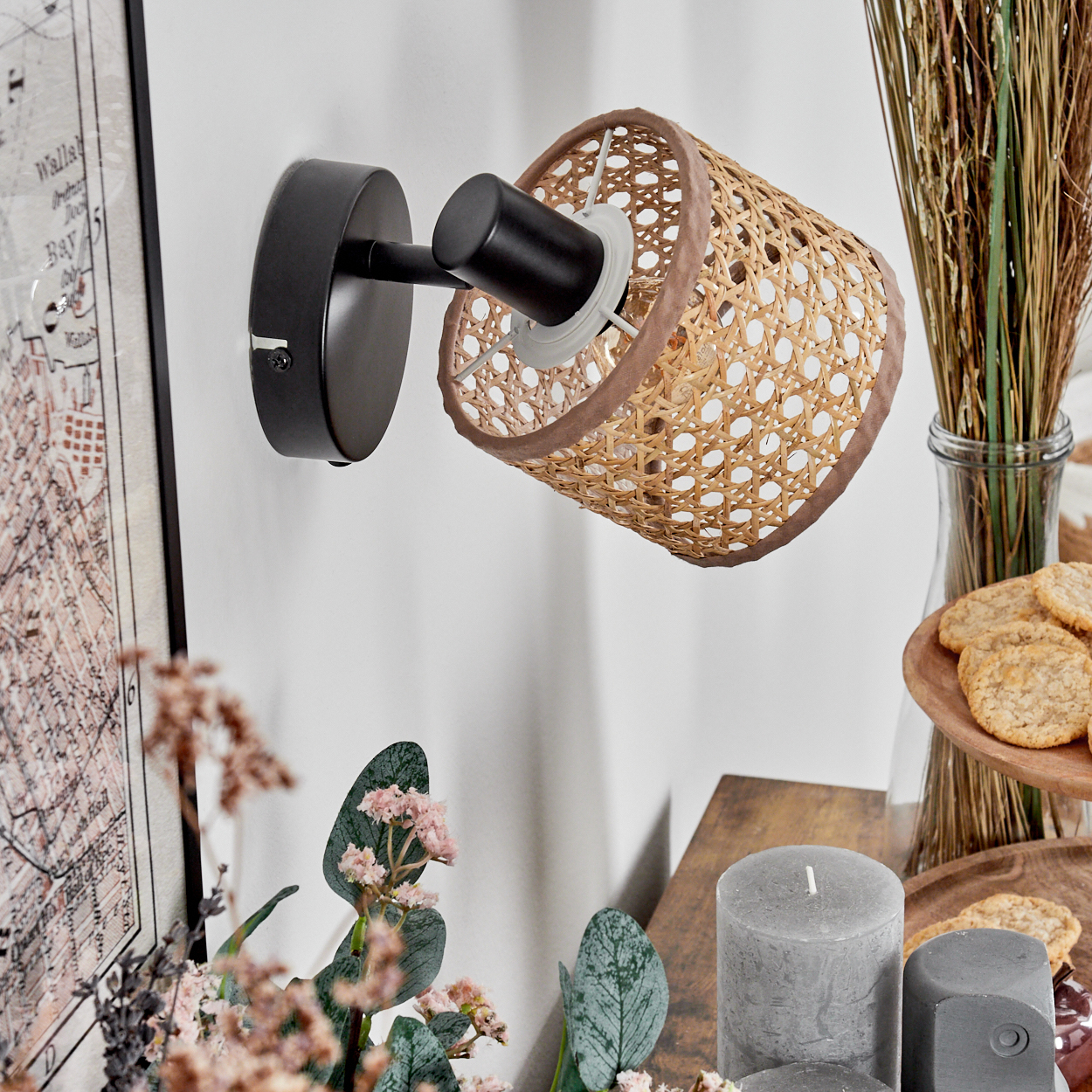 Eine Wandlampe mit gewebtem Lampenschirm ist an einer weißen Wand befestigt. Kekse, Kerzen und Blumen stehen auf einem Tisch.
