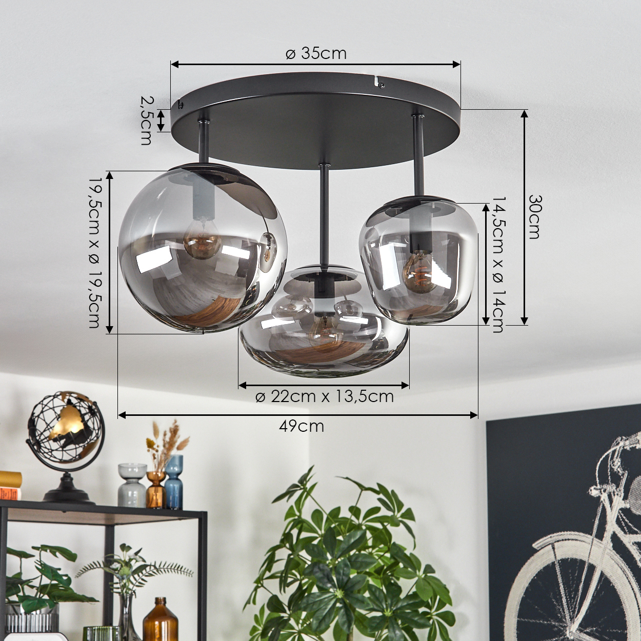 Eine moderne Deckenlampe mit drei Glasschirmen. Abmessungen sind mit Pfeilen angegeben. Raumdekor umfasst Pflanzen und ein Fahrradkunstwerk.