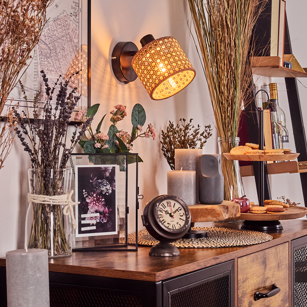 Eine gemütliche Innenszene mit Lampe, Uhr, Blumen, Kerzen und Snacks auf einem Holzschrank. Trockenblumen und eine Karte an der Wand.