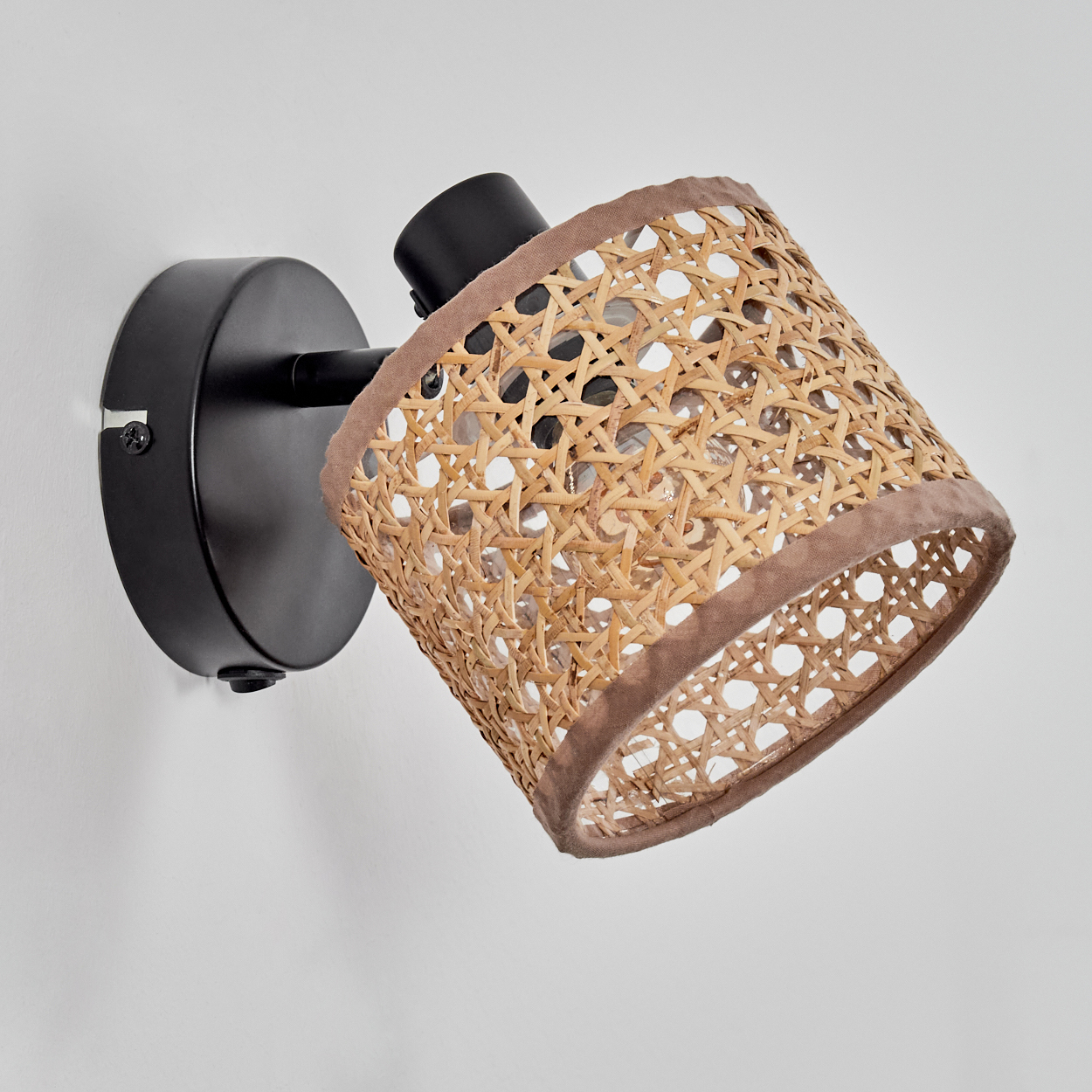 Eine Wandleuchte. Sie hat einen schwarzen Sockel und Arm mit einem Lampenschirm aus geflochtenem Rattan, der mit braunem Stoff verkleidet ist. Die Wand ist weiß.