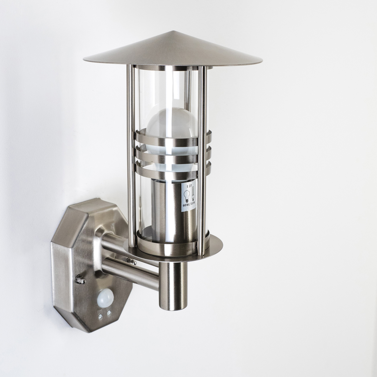 Eine silberne Außenlampe an einer weißen Wand. Die Lampe hat eine Glühbirne in einem Glaszylinder und einen Bewegungssensor.