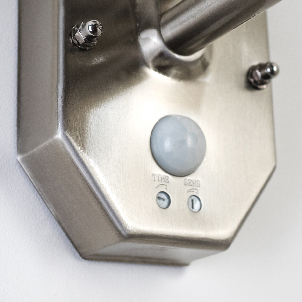 Ein silberner Bewegungssensor ist an einer Wand montiert. Er hat einen runden Sensor, Steuerungswahlschalter und eine silberne Metalloberfläche.