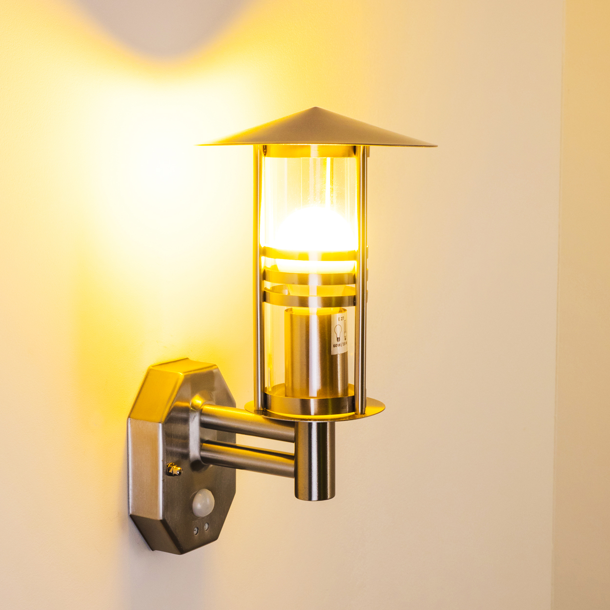 Eine an der Wand montierte Außenlampe leuchtet. Sie hat einen silbernen Körper, klares Glas und eine konische Oberseite. Die Wand ist weiß und das Licht ist gelb.