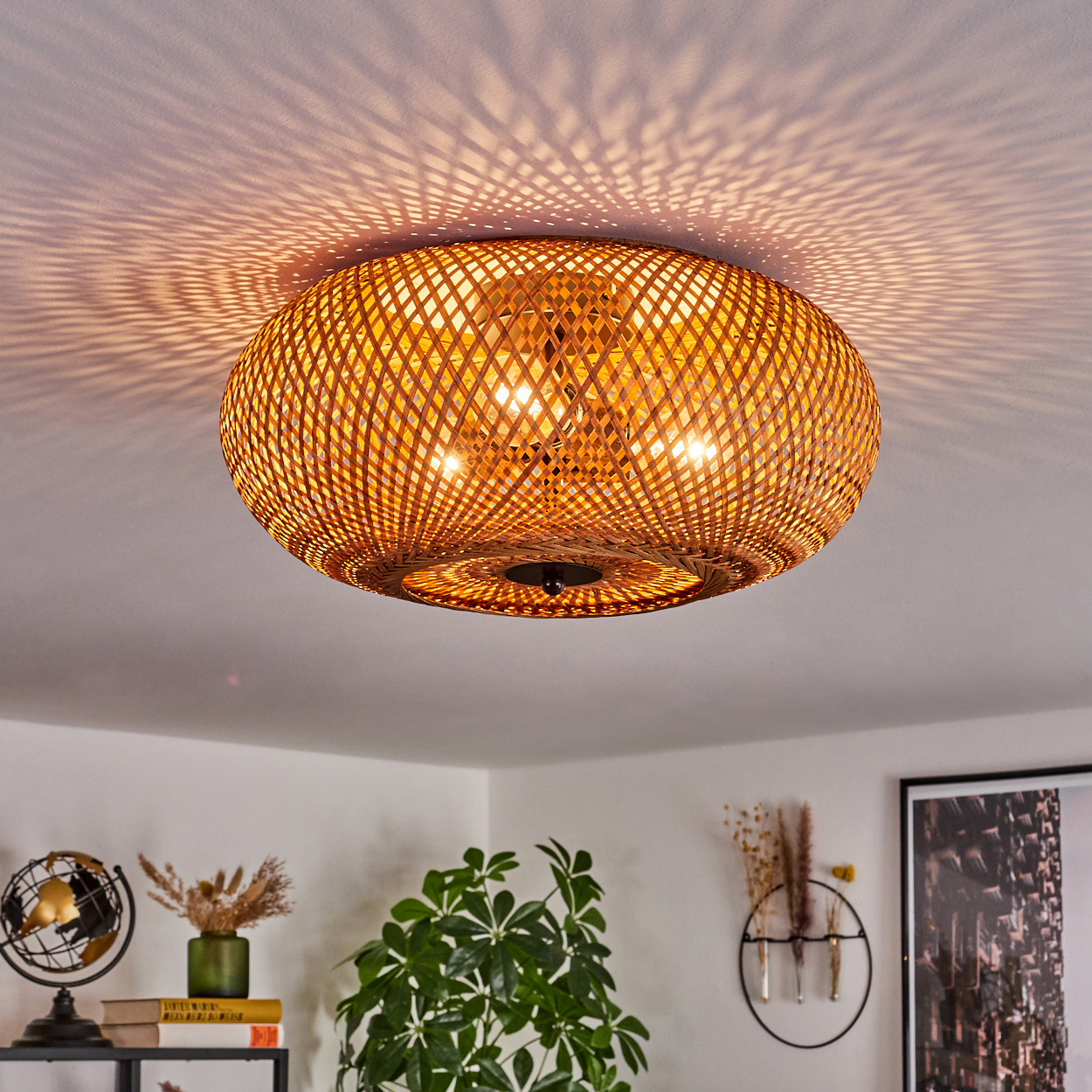 Eine Deckenlampe aus gewebtem Bambus hängt an einer weißen Decke und wirft Schattenmuster. Pflanzen, ein Globus und Deko sind zu sehen.